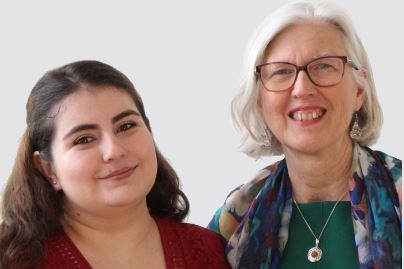 [ai] Two women standing side by side, smiling at the camera. The woman on the left has long dark hair and is wearing a red top. The woman on the right has gray hair, glasses, and is dressed in a green top with a colorful scarf.
